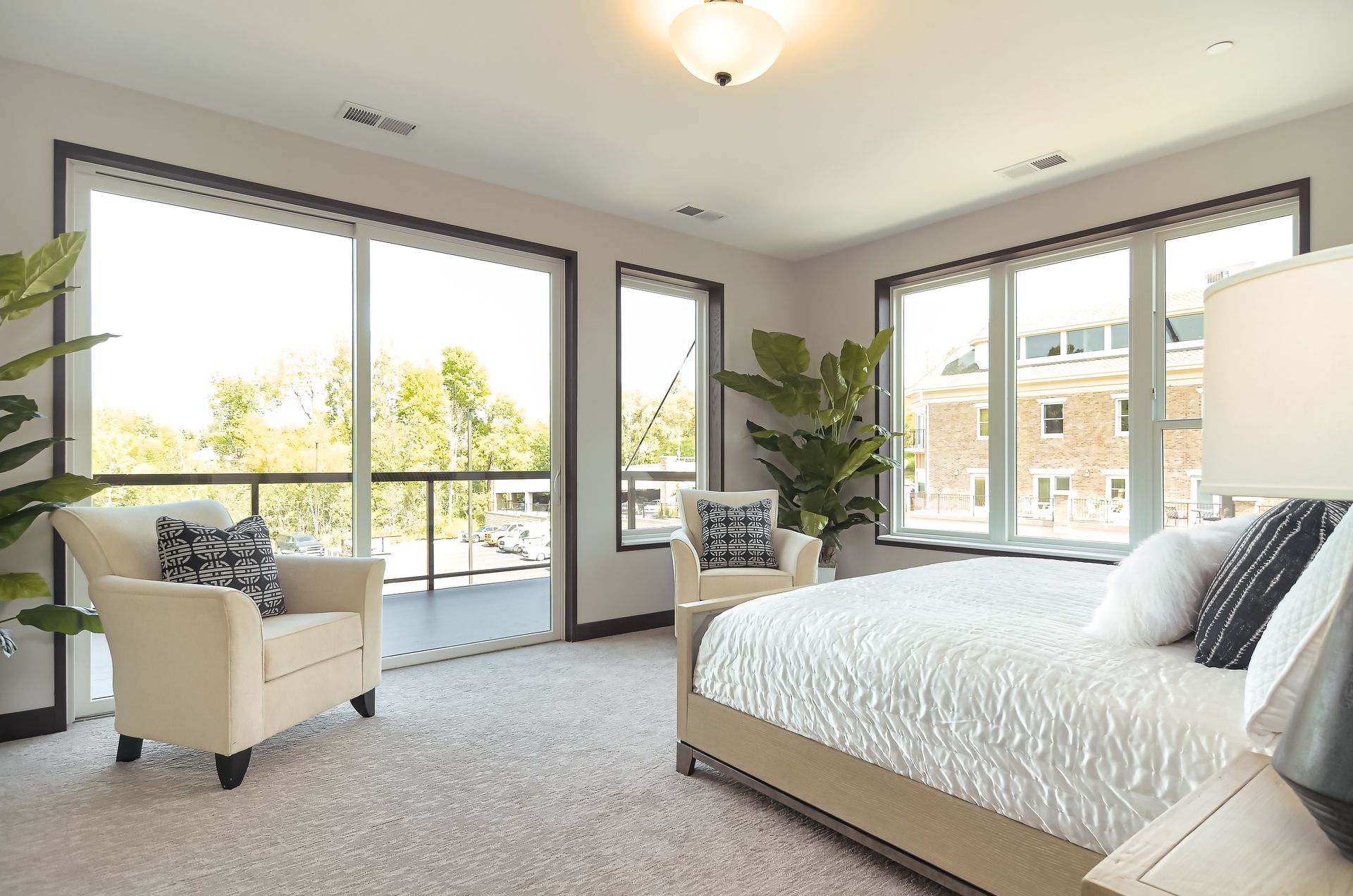 Bright bedroom with large windows, cream-colored armchair, and a bed with white bedding.