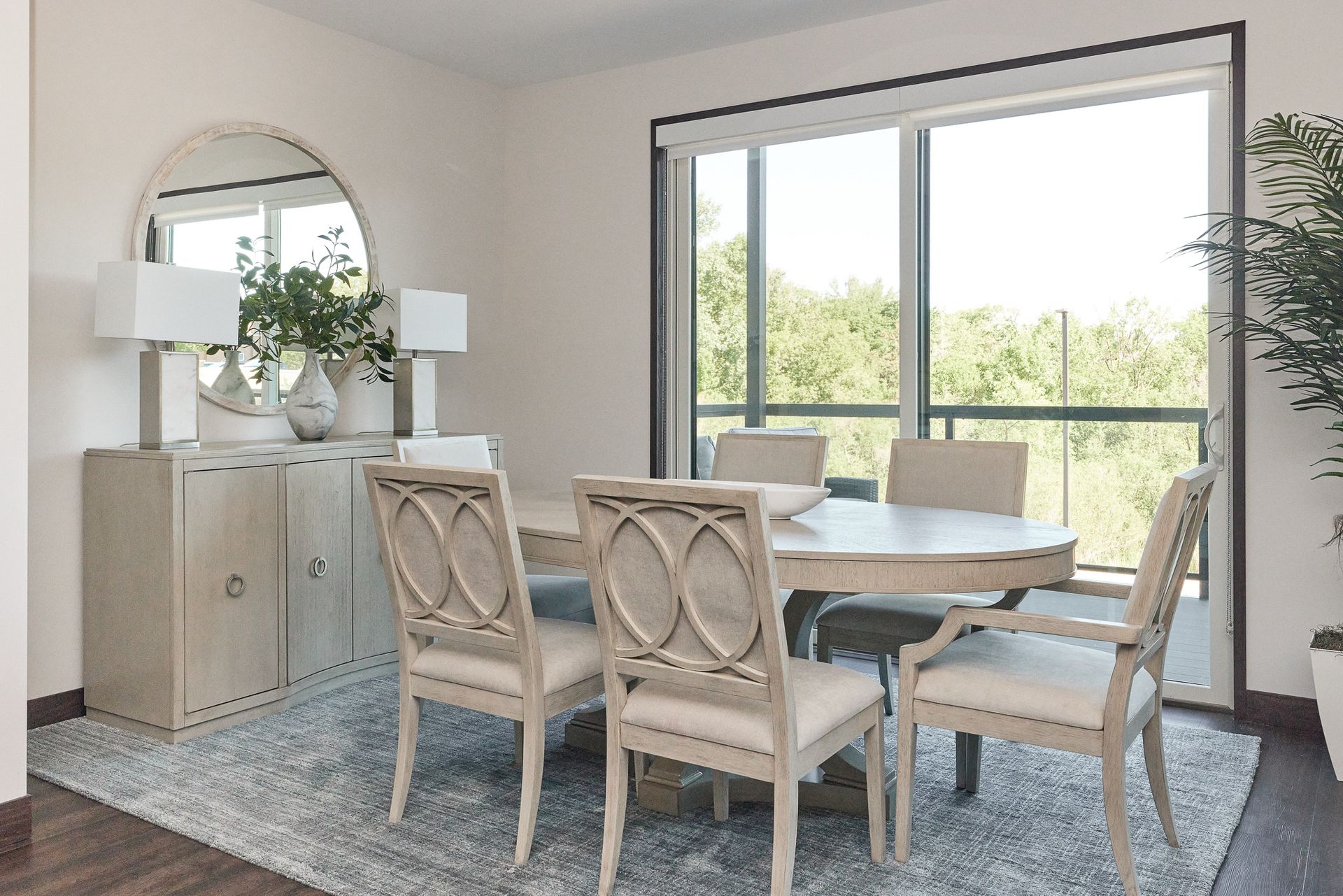 Dining room with round table, six chairs, buffet with mirror, rug, and large window with view.