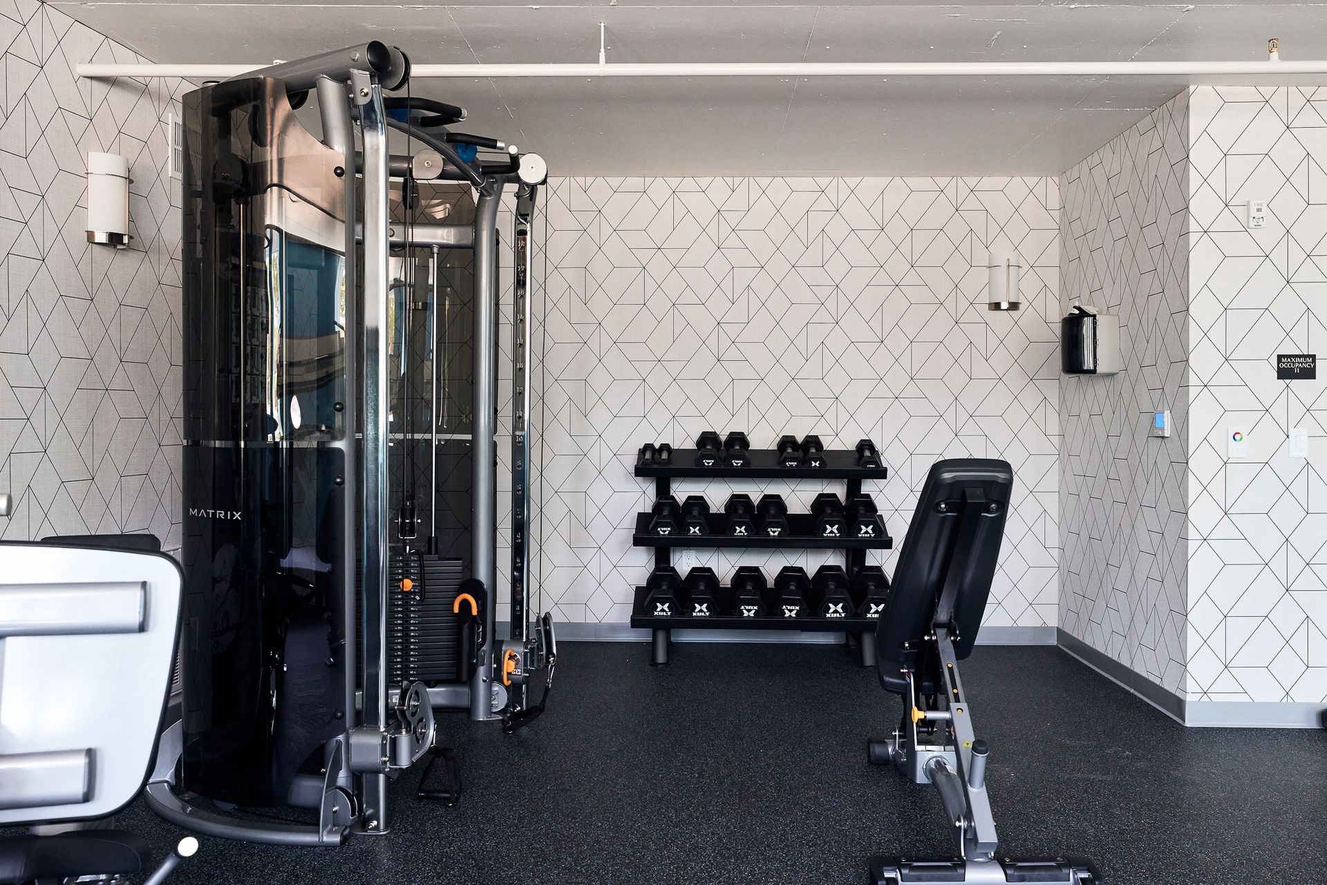 Gym interior with weight machine, weights, and bench on black flooring, white patterned walls.
