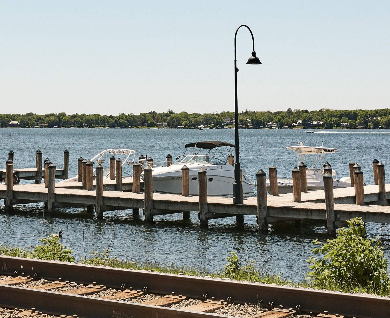 Wooden pier with parked boat, lamp post, and water, with trees on the horizon. Railroad tracks in foreground.