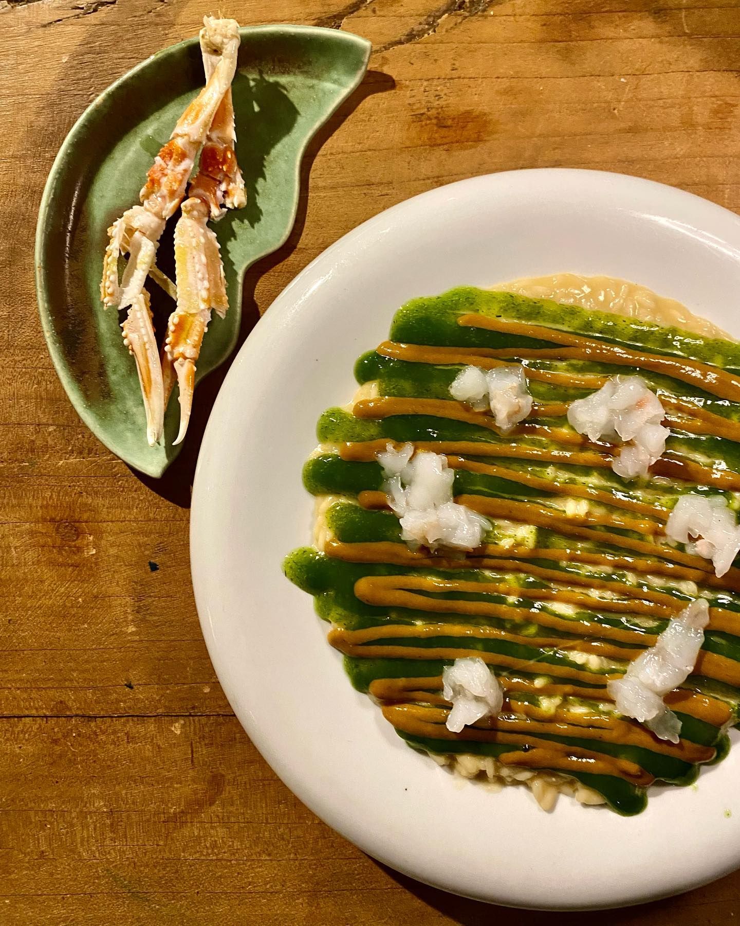 Un piatto bianco condito con salsa verde accanto a un piatto di gamberi su un tavolo di legno.