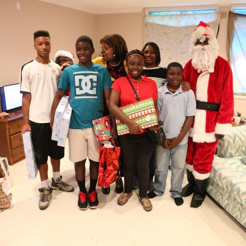 Group of people and Santa Claus pose indoors with gifts.