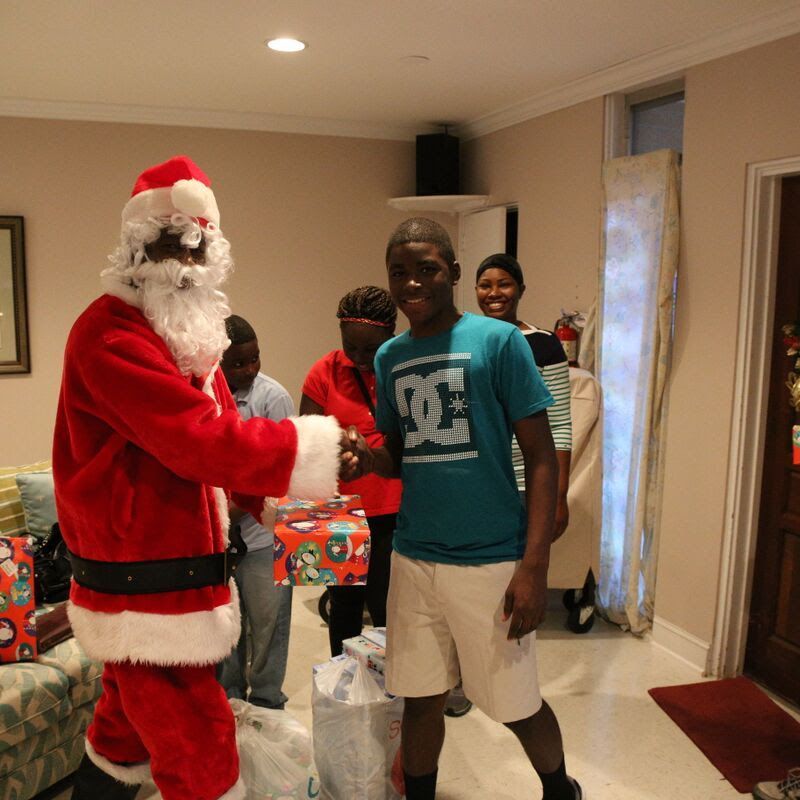 Santa shakes hands with a young Black man, handing him a wrapped gift. Indoors, others watch.