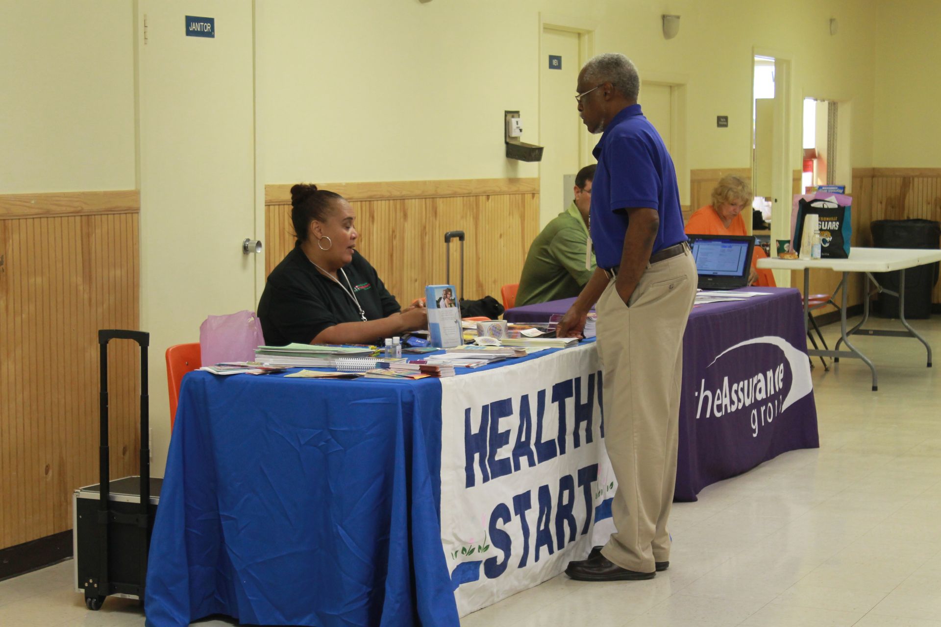 A woman at a health information table speaks with a man. Interior setting with other people.