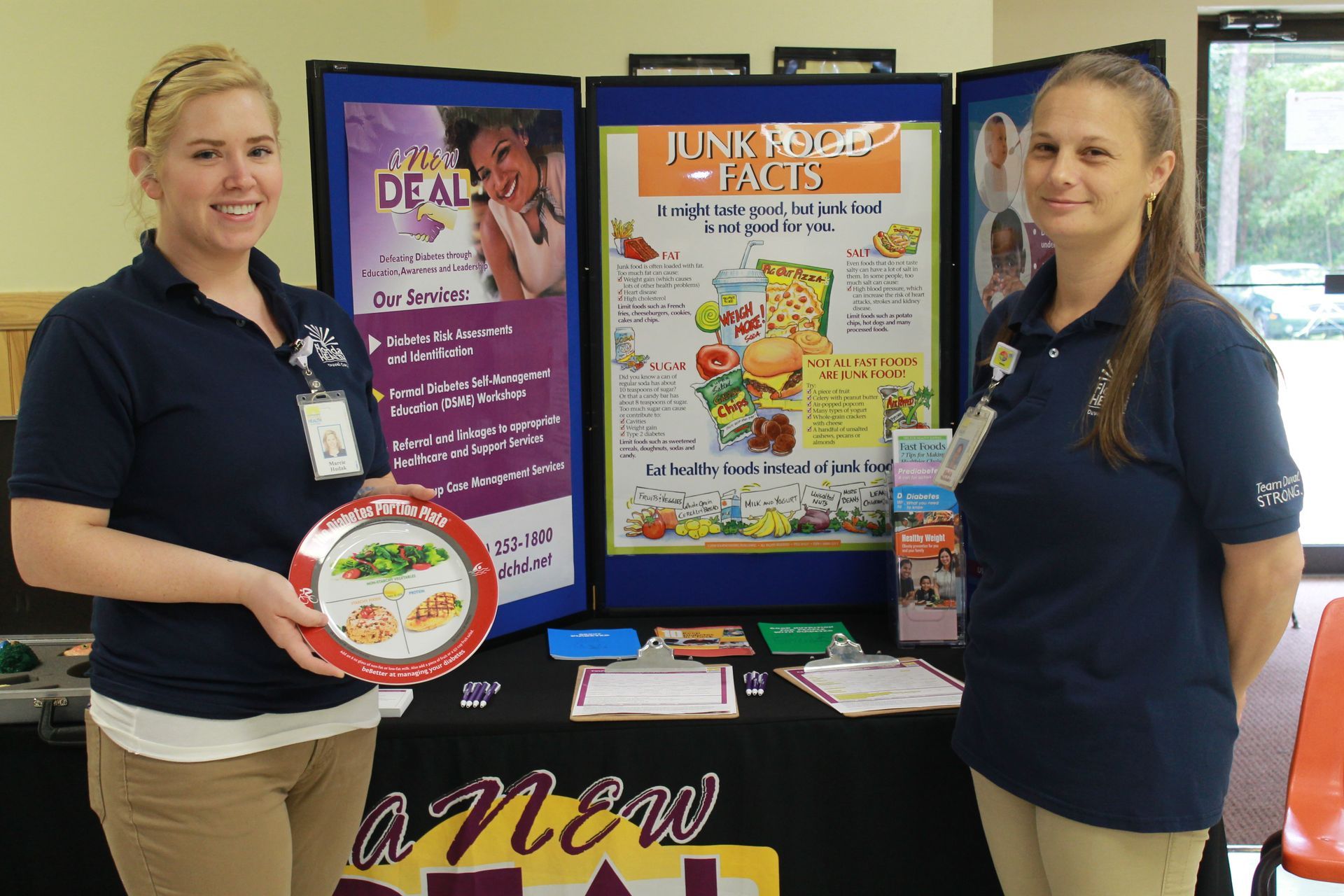 Two women at a health fair, stand by a table with nutrition posters. One holds a plate.