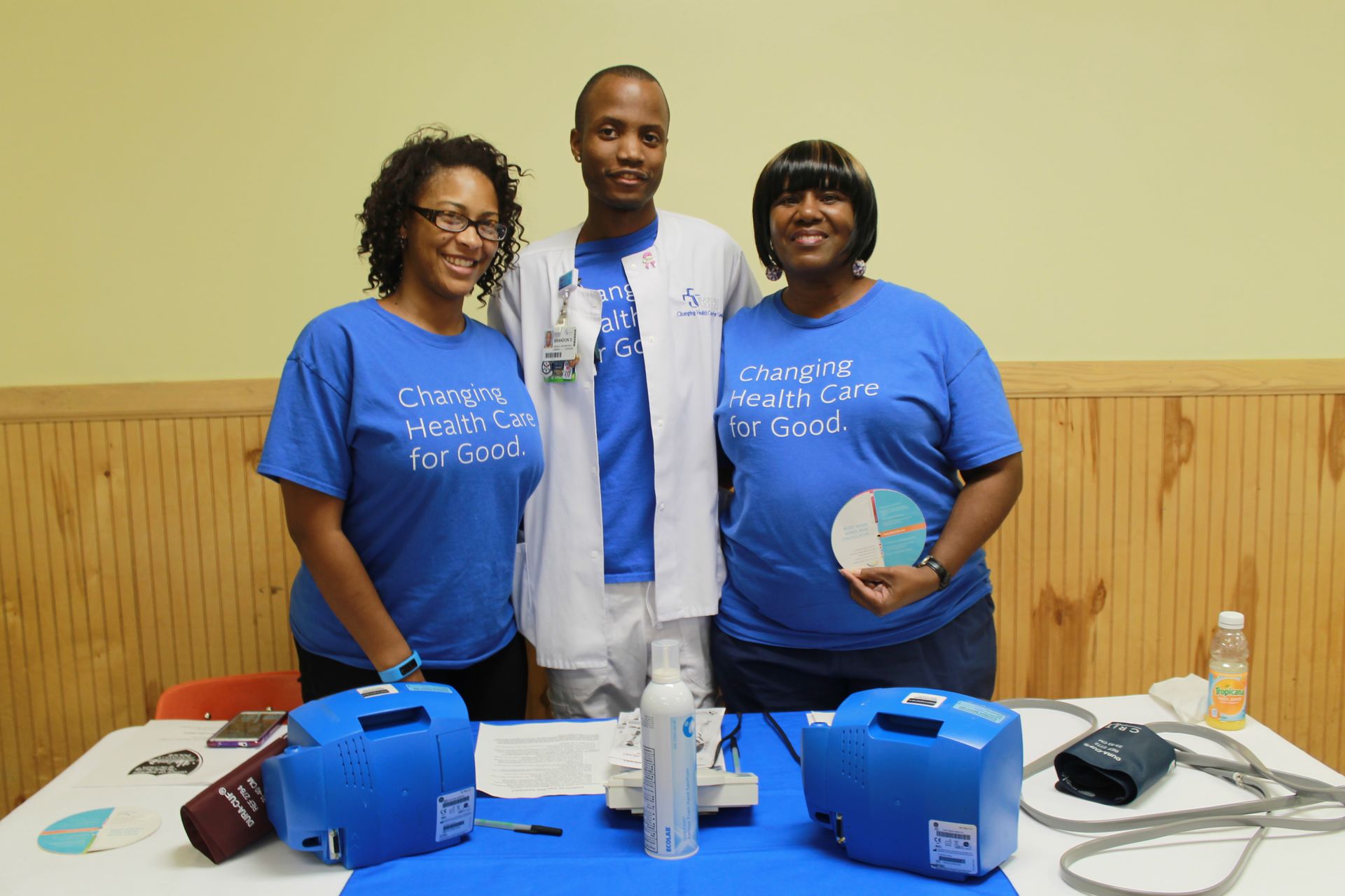 Three healthcare workers at a health fair, wearing blue shirts. Two women and a man stand behind a table.