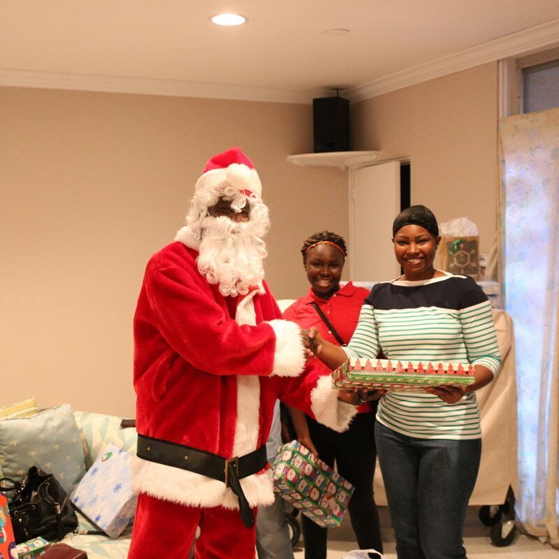 Santa Claus giving a gift to a woman in a striped shirt. Two women smile in a living room.