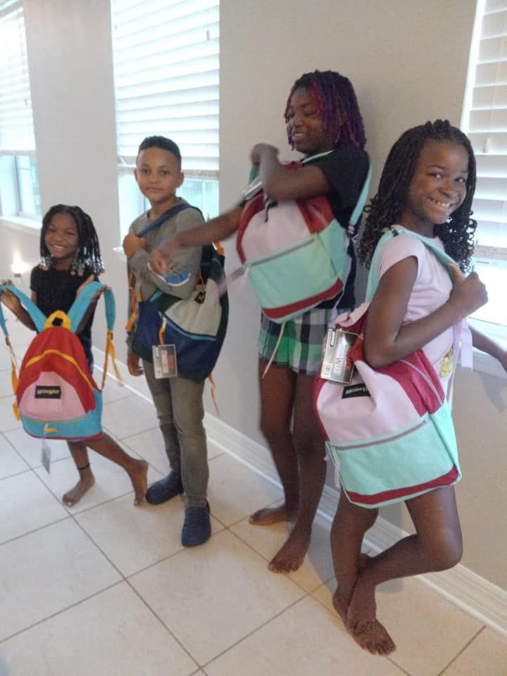 Four Black children pose with colorful backpacks indoors, smiling.