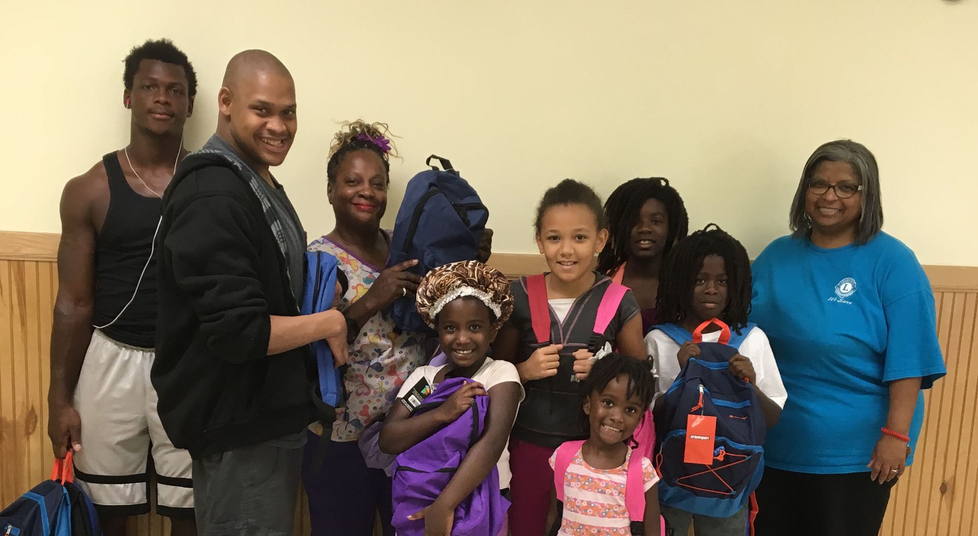 Group of people, mostly Black children and adults, holding backpacks, indoors, smiling.