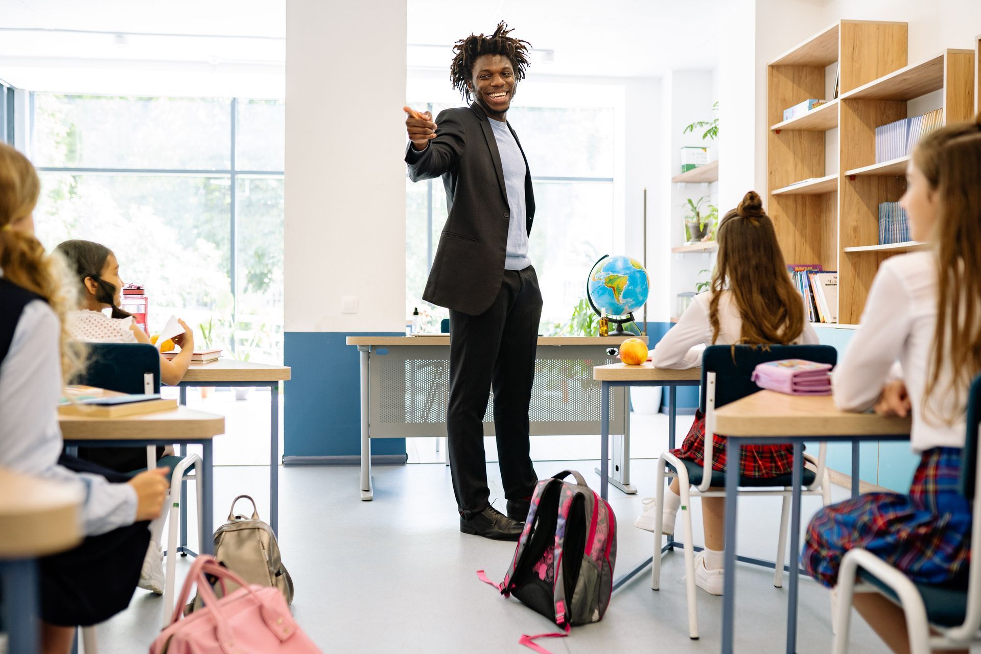Teacher stands in front of students in a classroom, gesturing; a globe and desk are visible.