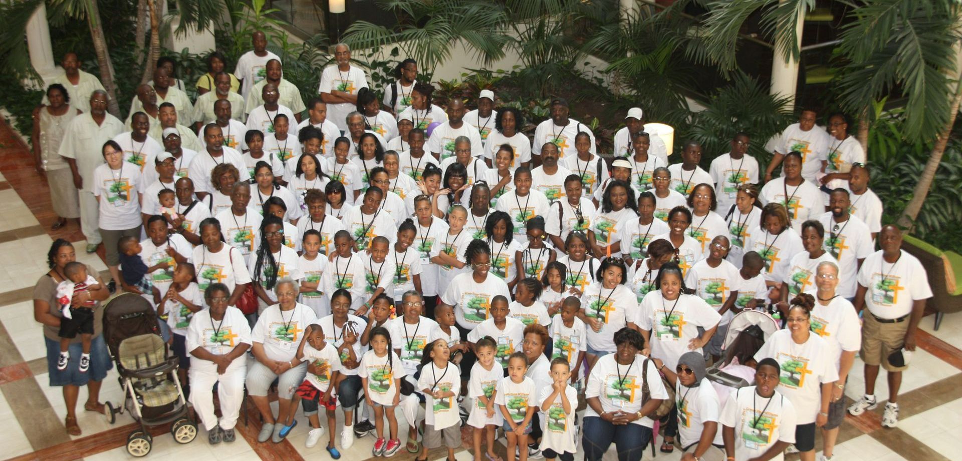 Large group of people in white t-shirts posing for a photo, outside, greenery in the background.