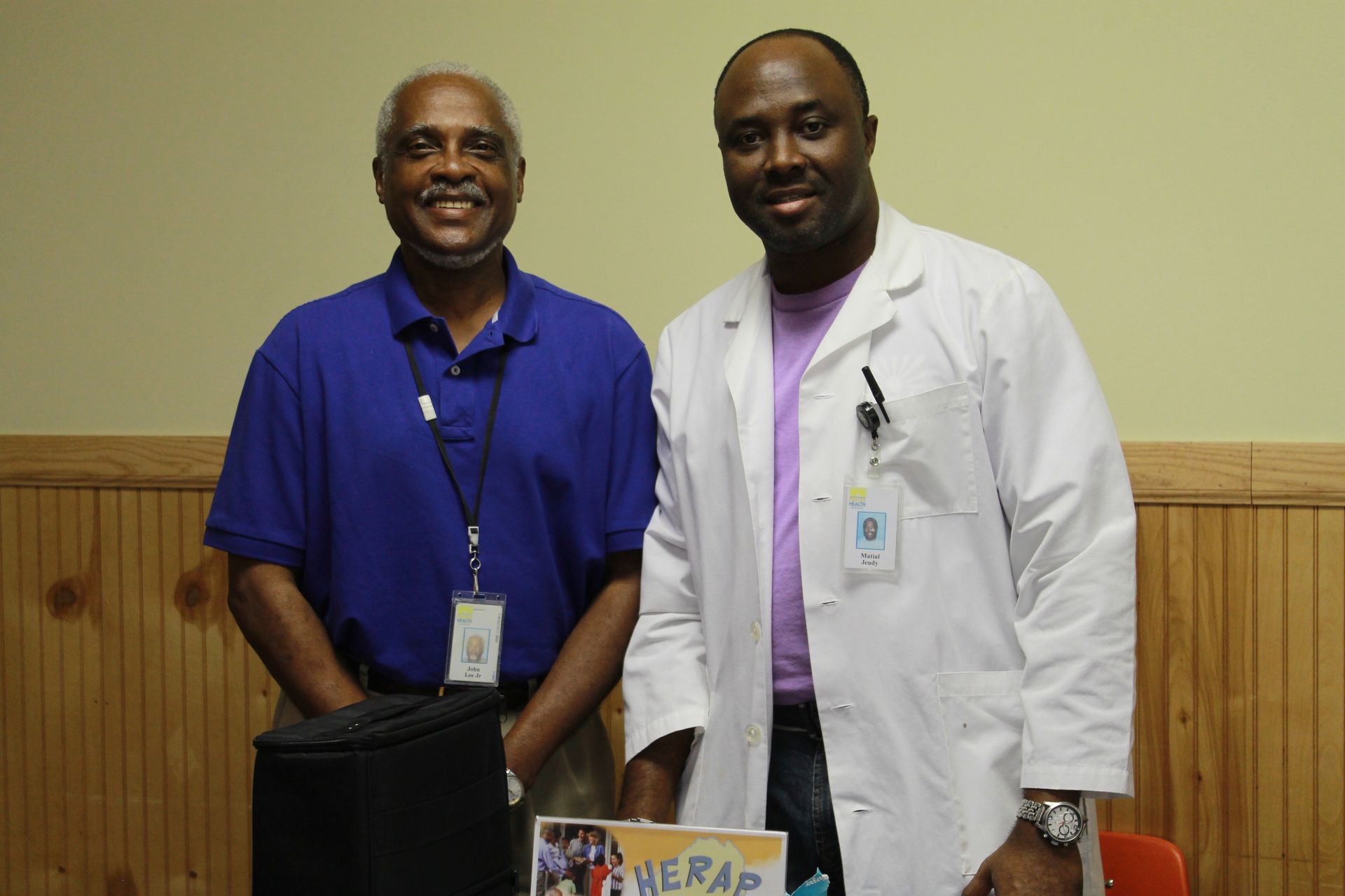 Two Black men smiling at a table. One in a blue shirt, the other in a lab coat. Interior.