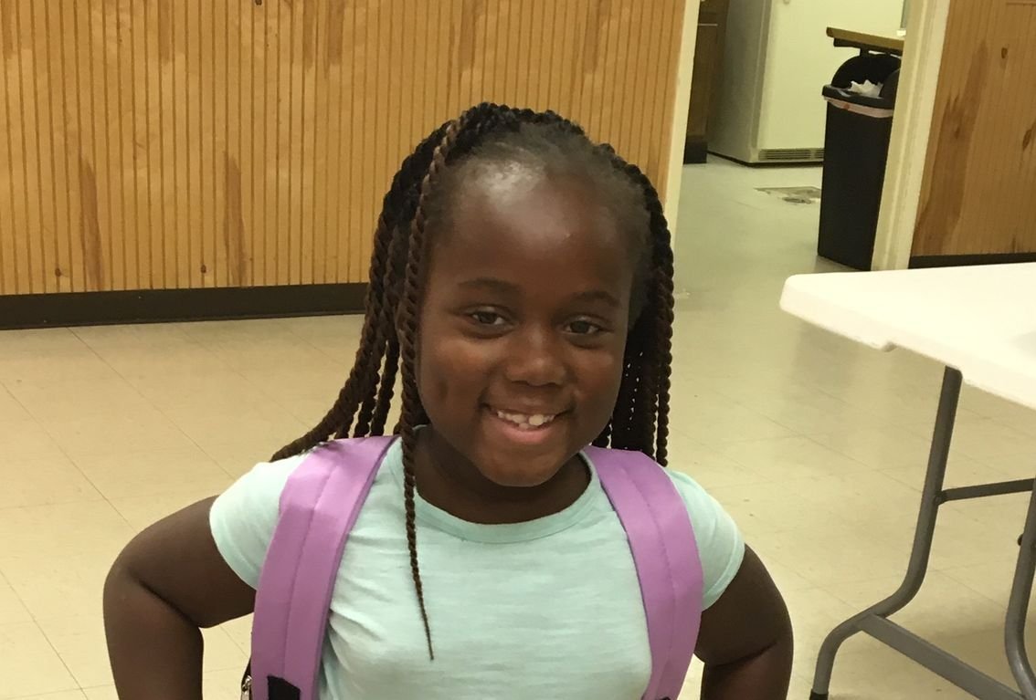 Young Black girl with braids and purple backpack smiles at the camera, indoors.
