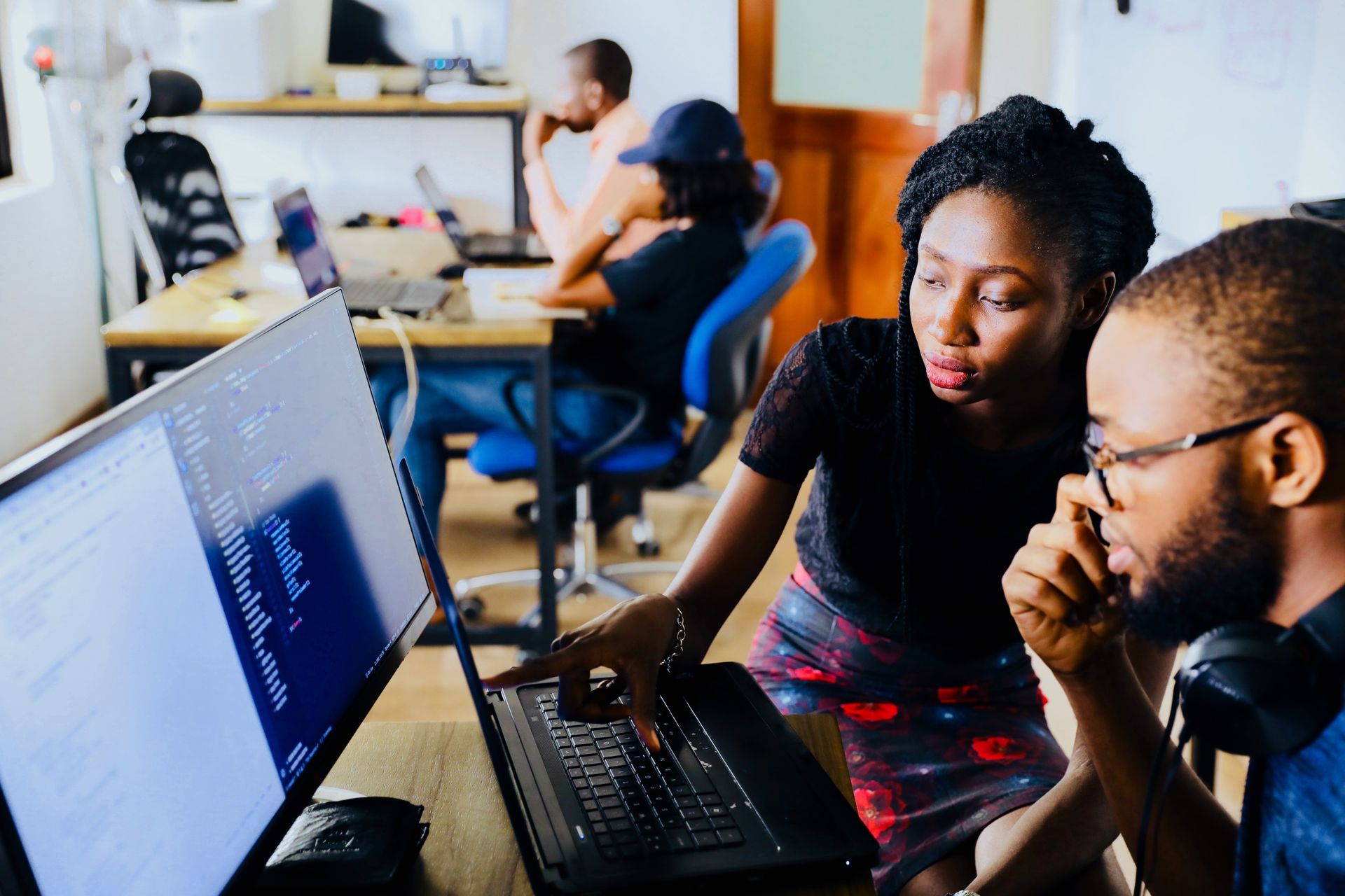 Two people looking at computer code, in an office setting. Woman points at laptop screen as man watches. Other colleagues.
