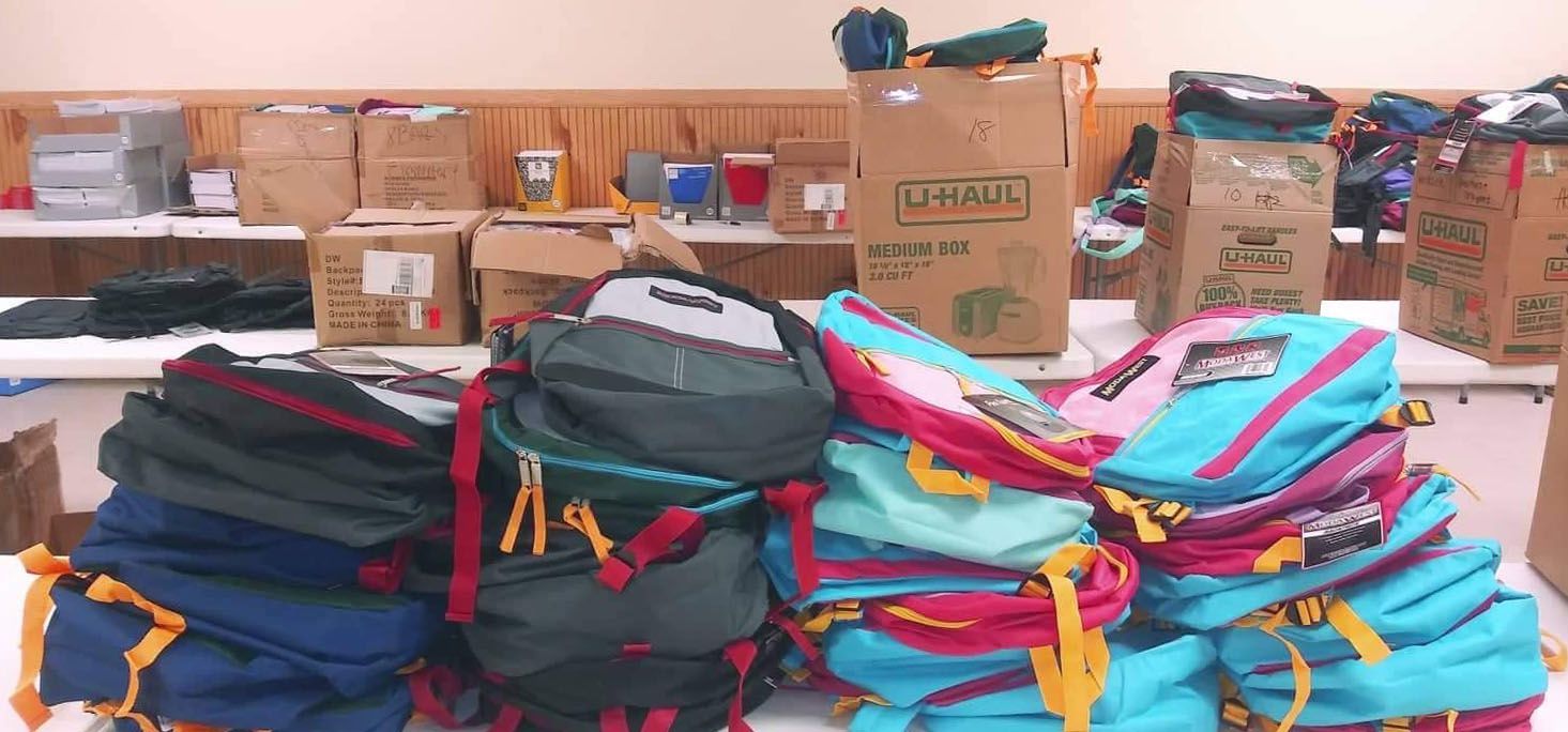 Stacks of colorful backpacks and boxes of school supplies on a table.