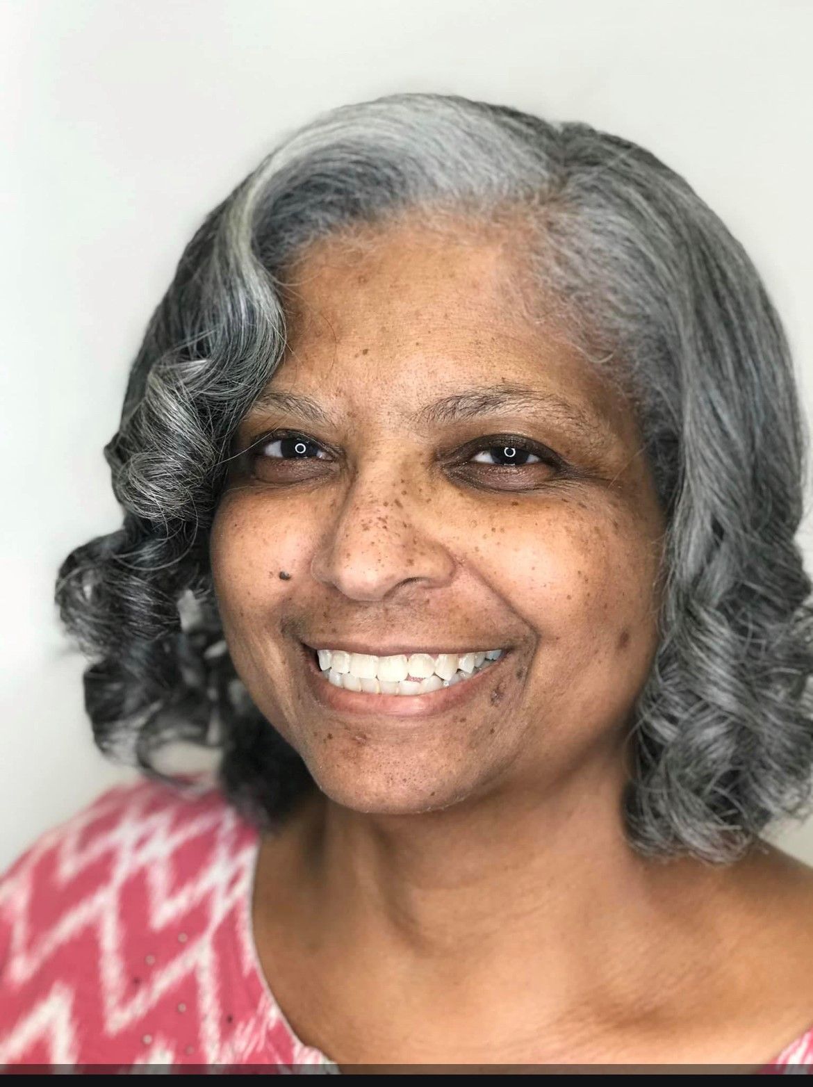 Smiling Black woman with gray curly hair, wearing a pink patterned top.