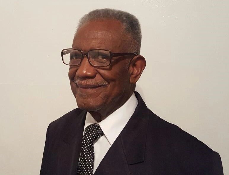 Older Black man wearing glasses, smiling, in a dark suit and tie, white shirt.