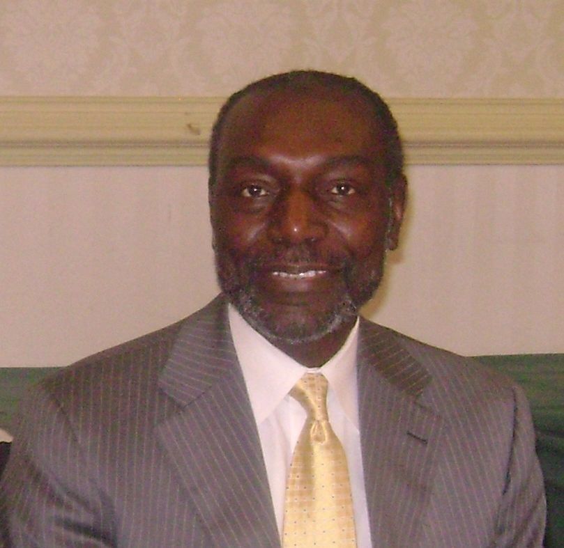 A Black man in a suit smiles at the camera. The setting appears to be an office or meeting room.