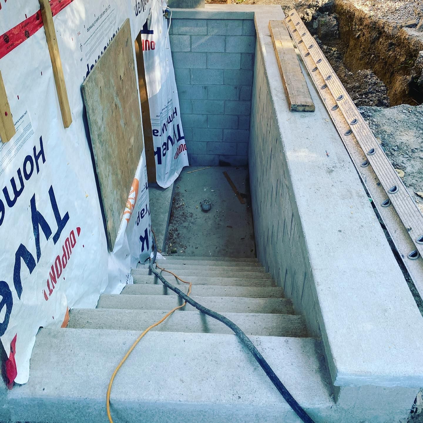 Concrete basement stairs leading downwards, construction site, Tyvek wrap, gray brick wall, wooden boards, ladder.