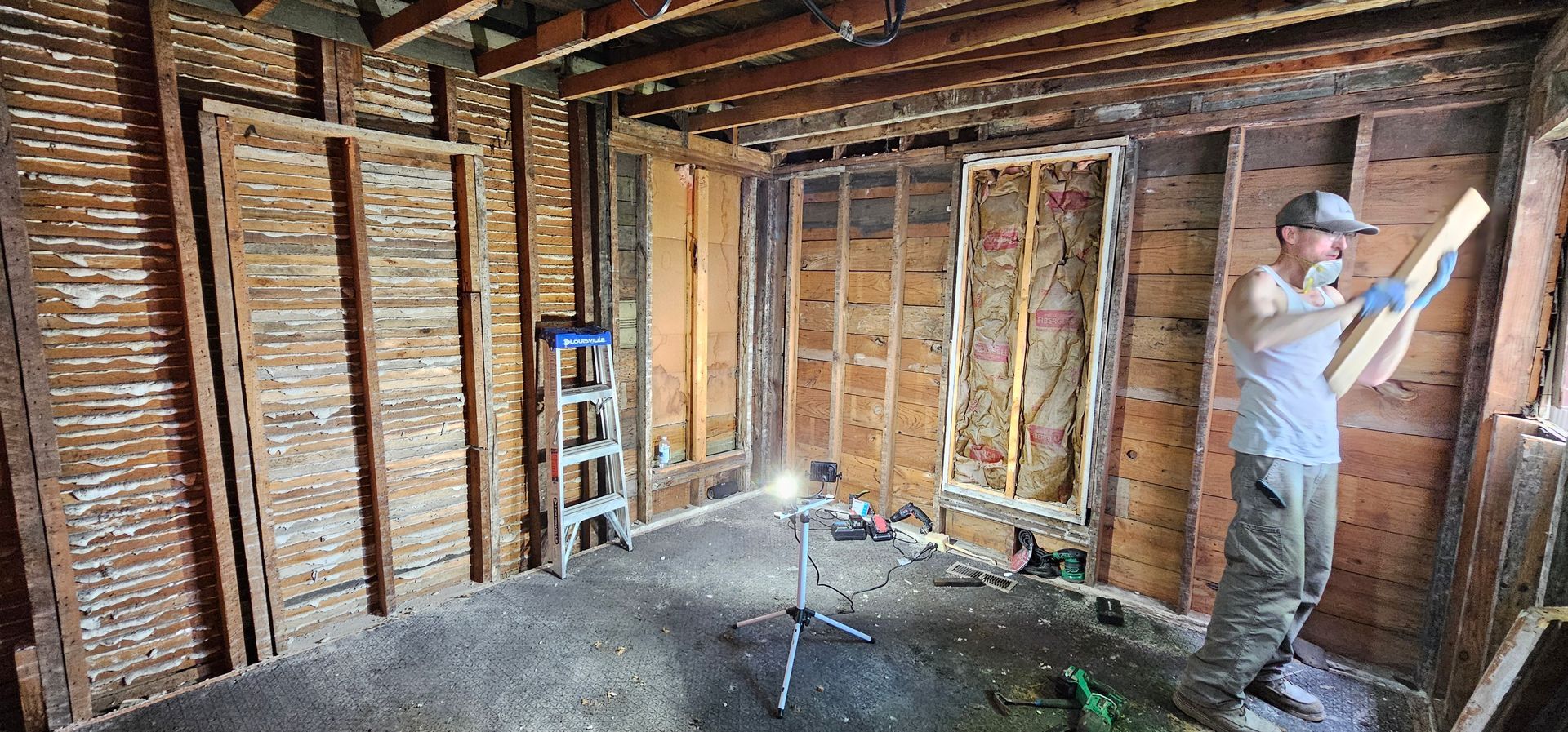 Empty basement with exposed beams, concrete floor, wooden stairs, and a lit work area.