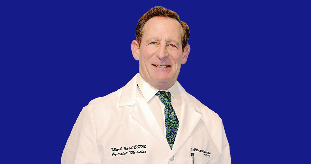 A man in a white lab coat and tie is smiling against a blue background.