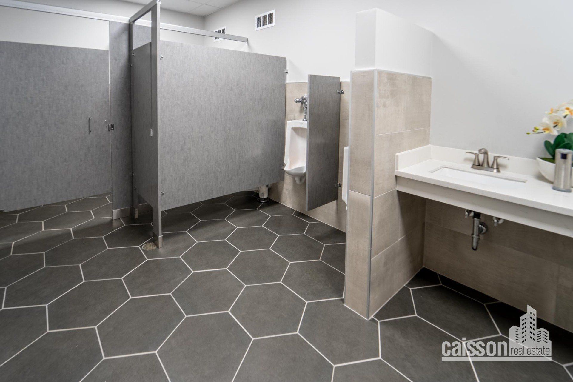 Interior view of modern restroom with grey flooring, grey stalls, and flowers on sink.
