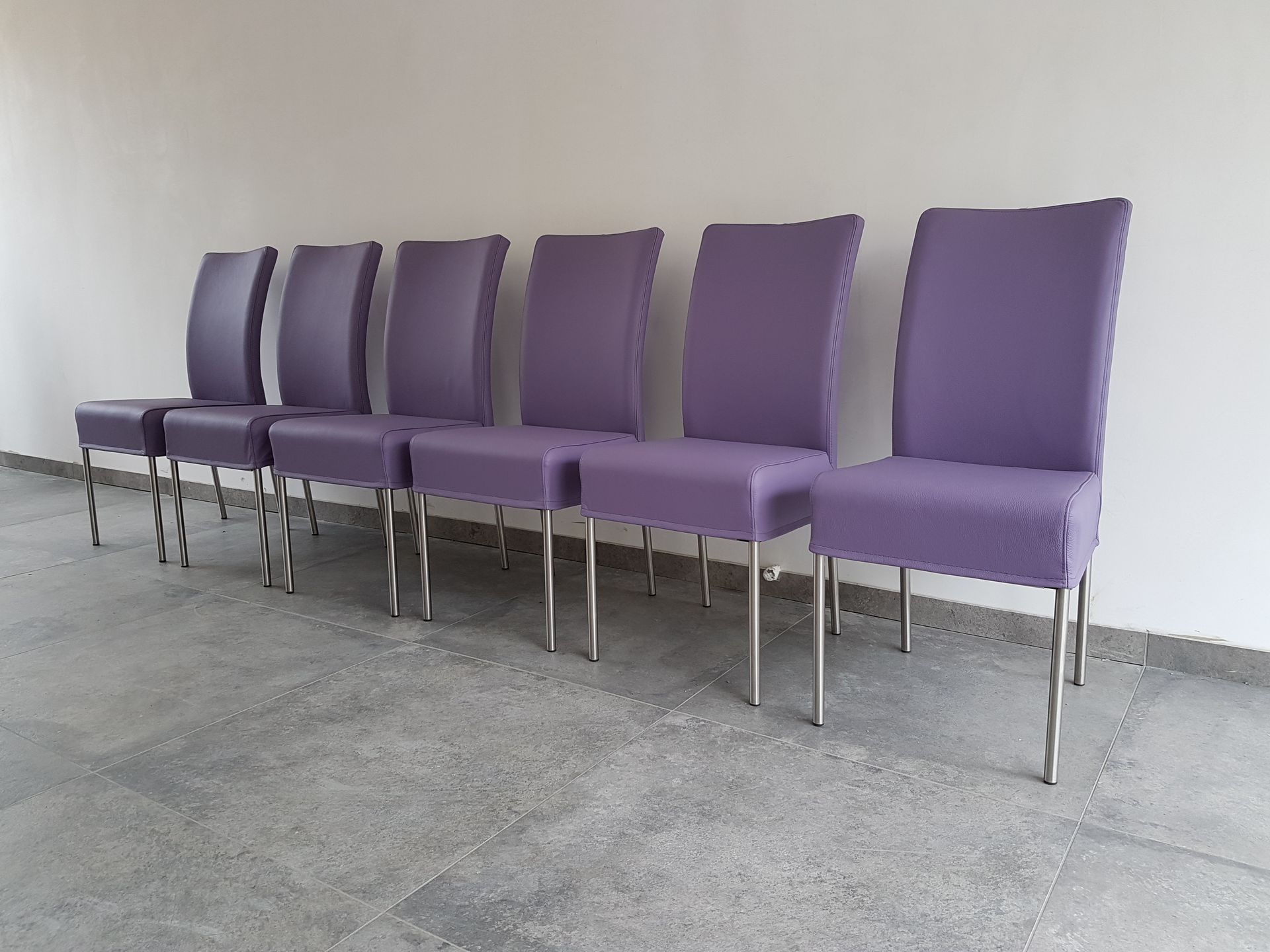 Een rij van zes moderne, paars gestoffeerde stoelen met metalen poten staat tegen een effen witte muur.