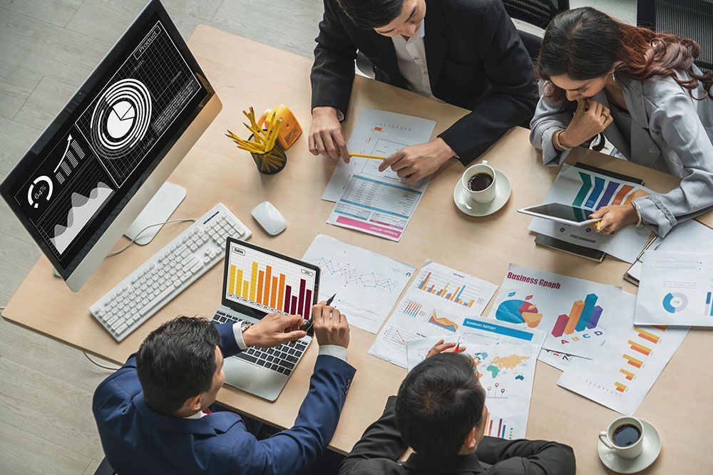 Four colleagues collaborate at a wooden table reviewing business charts and data on computer screens and printed documents.
