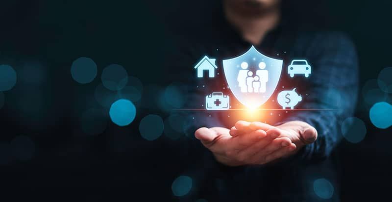 Hands holding a glowing shield icon containing family, home, car, health, and money symbols against a dark background.