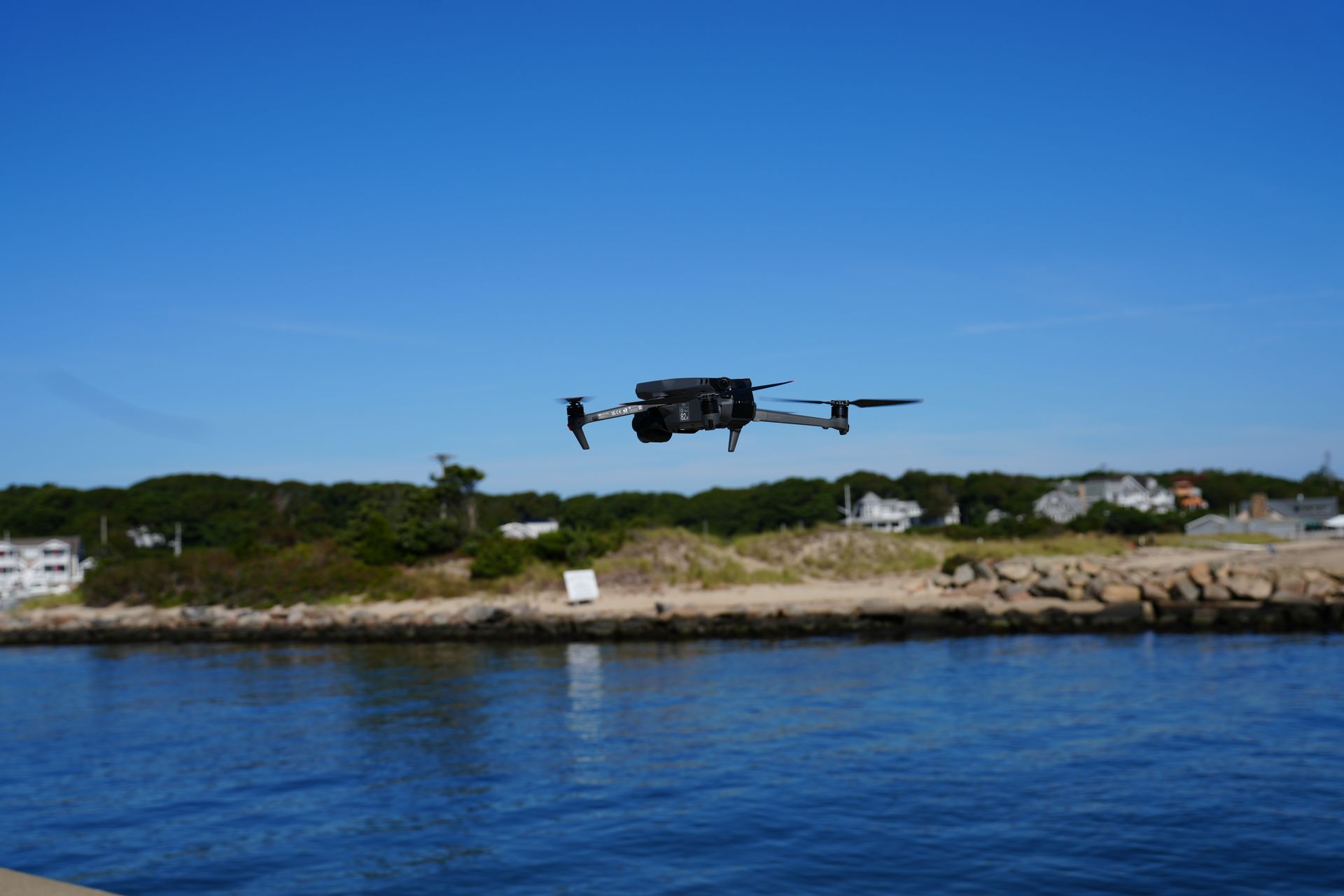 Drone flying over water with a shoreline and blue sky in the background.