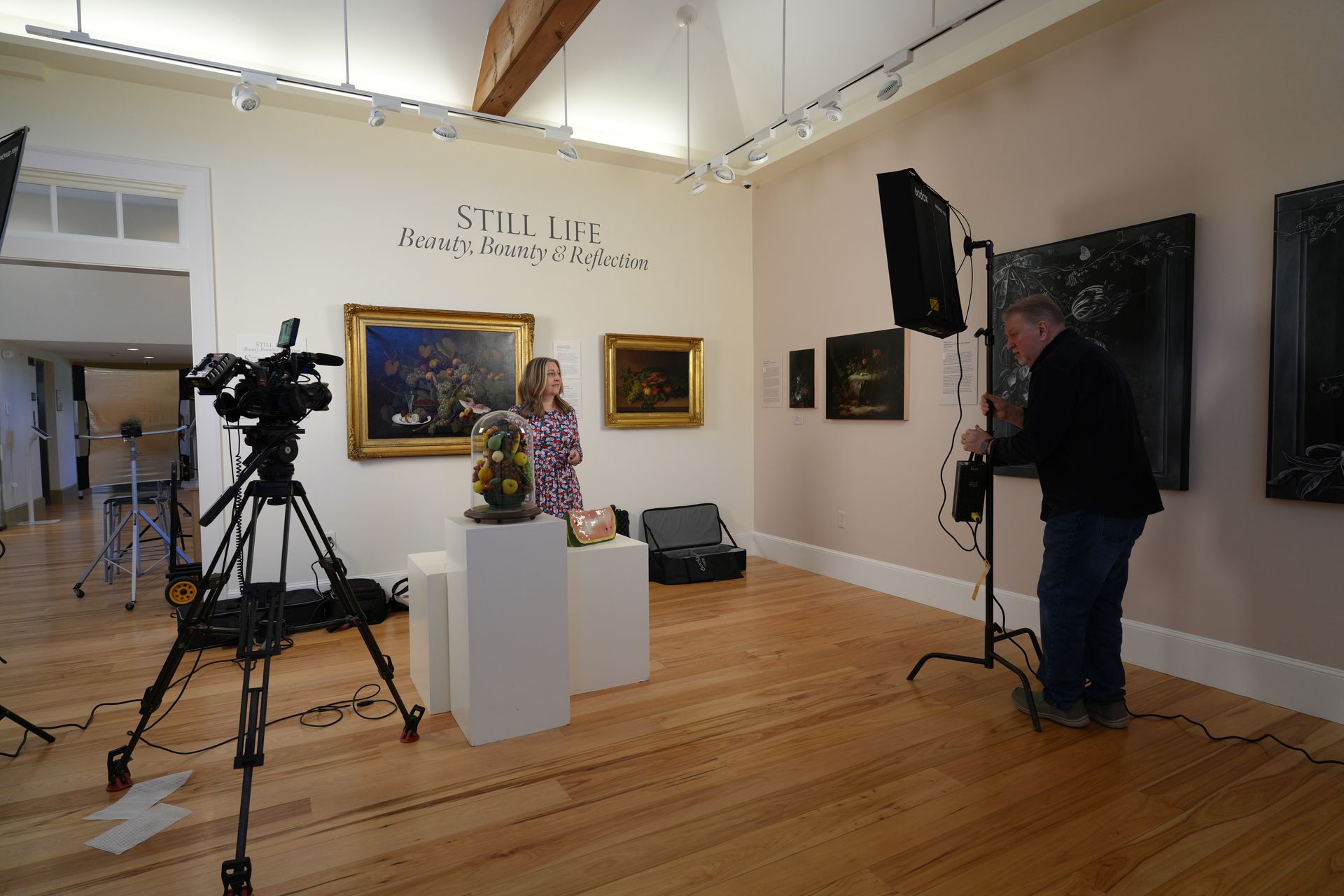 Inside a gallery, a woman stands before an artwork, a cameraman adjusts equipment; filming in progress.