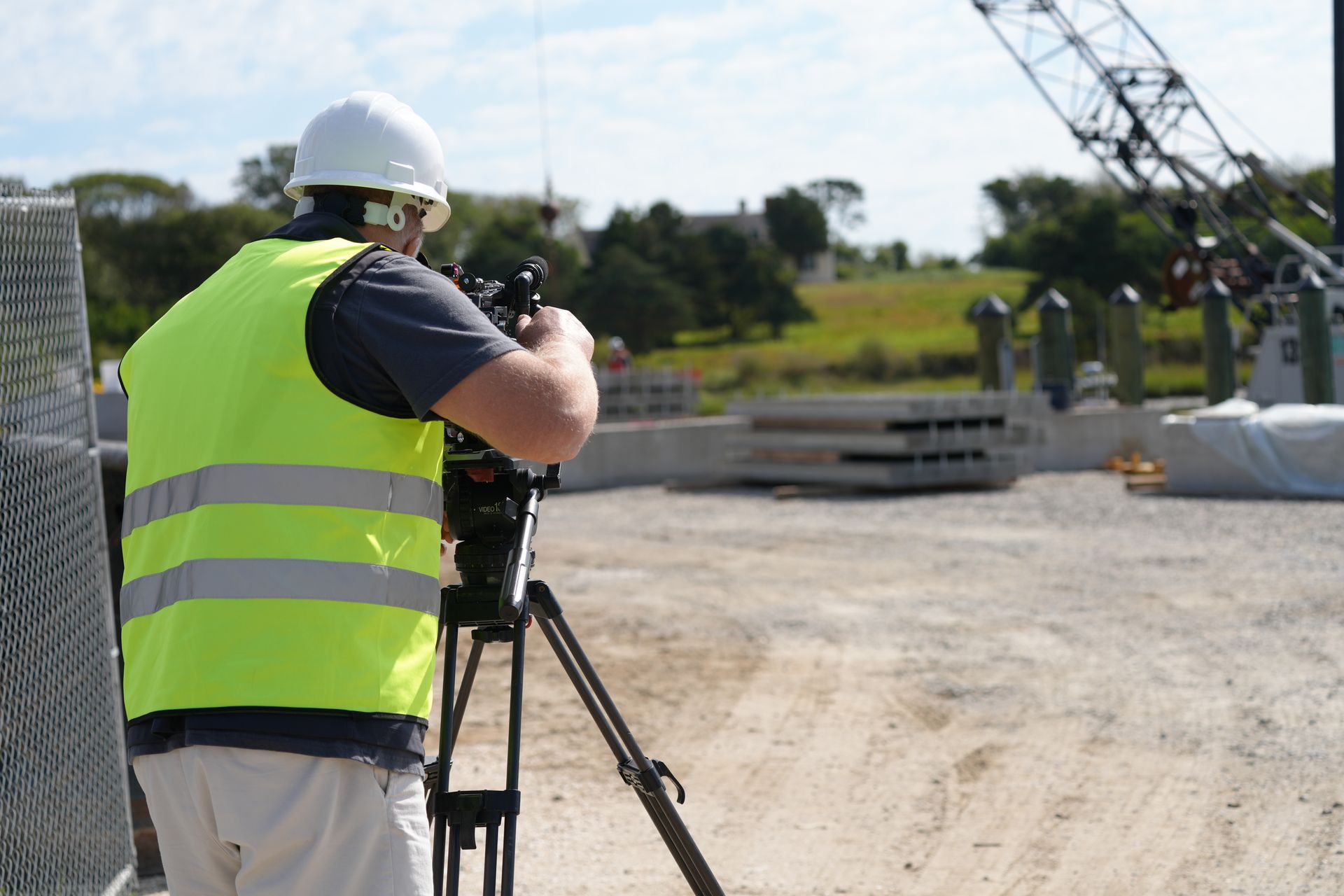 Construction worker filming with a camera on a tripod, wearing a white helmet and reflective vest.
