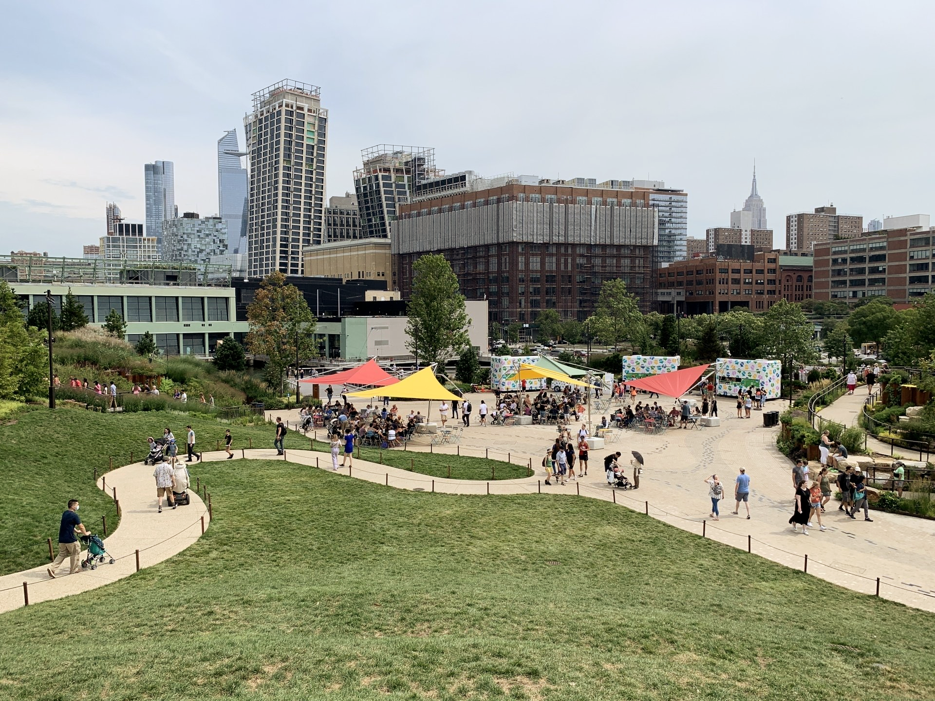 New York City skyline is in the background with the Edge Observatory and Empire State Building prominent.  In the forefront is the dining area with 3 food trucks, yellow & red bright coverings for the seated areas with plenty of people seated and walking around. Closer to the forefront on the left is the winding, accessible pathway.