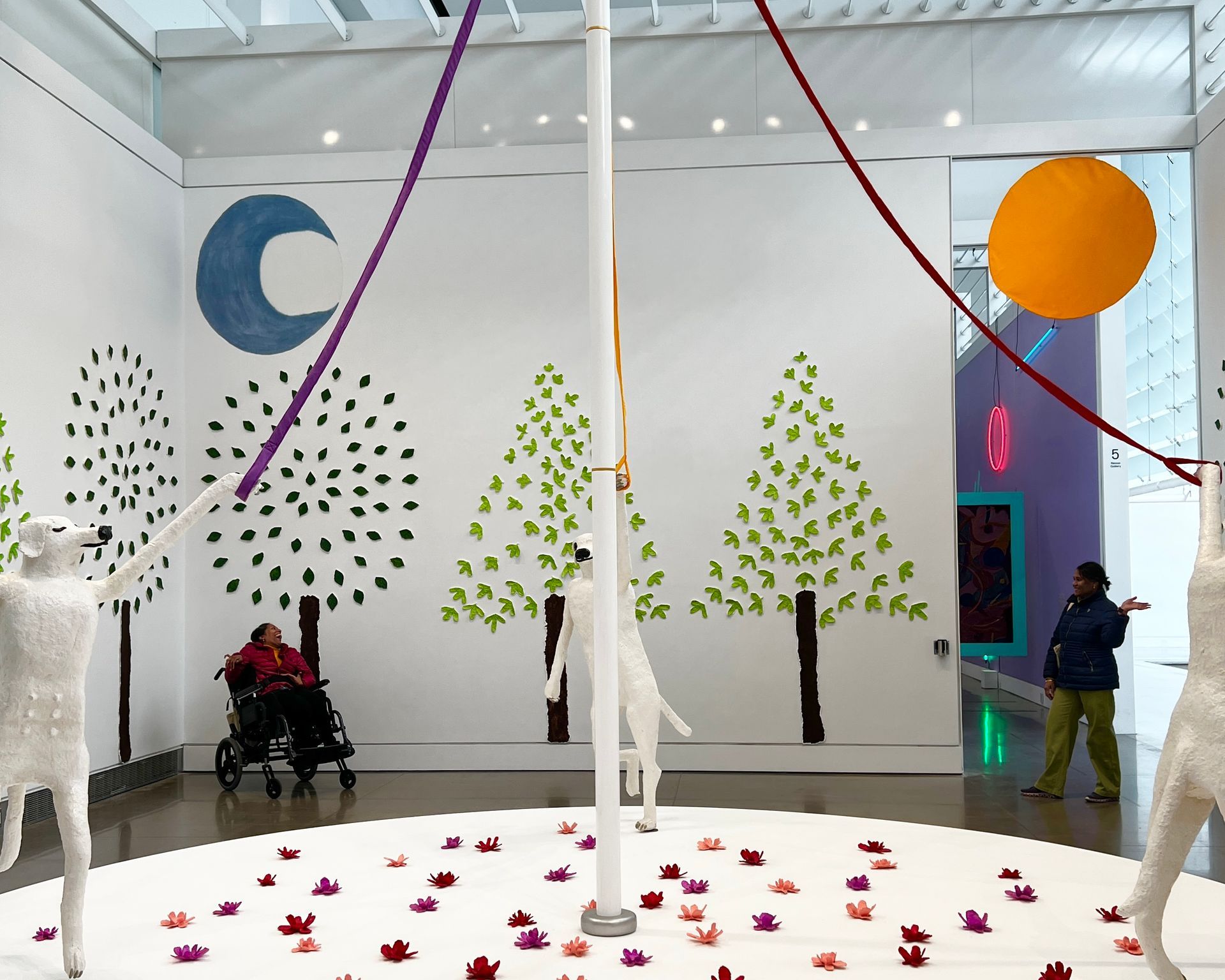 In the photo we took, the art installation sits within a bright, modern gallery space. At the center is a white circular platform adorned with numerous colorful flowers arrayed in shades of red, pink, and burgundy. Long ribbons in purple and red drape from the ceiling, converging on a single white pole—a symbolic white cane—at the circle's center. Three white dog figures, sculpted in a human-like form, are positioned around the platform. They interact with the pole and ribbons in what appears to be a dynamic and possibly celebratory fashion, injecting life into the scene. The wall art around the installation features stylized trees with leaves in shades of dark and light green, a blue crescent moon, and a full orange circle representing the sun. The ambiance is one of serenity, with the installation drawing viewers into its playful and colorful space.