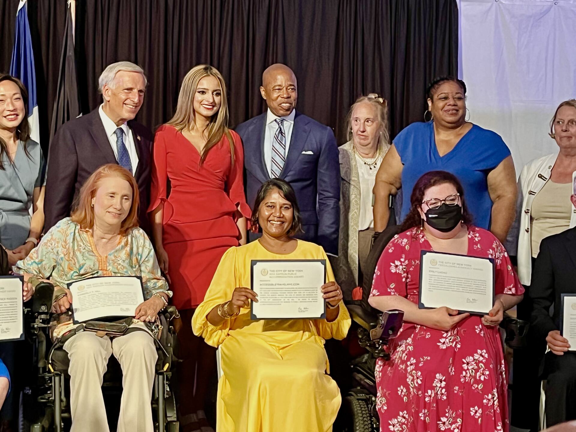 On a stage, three of the awardees, Kim Hill, Emily Ladau and myself holding our certificates. There are several government officials behind us including Mayor Adams.
