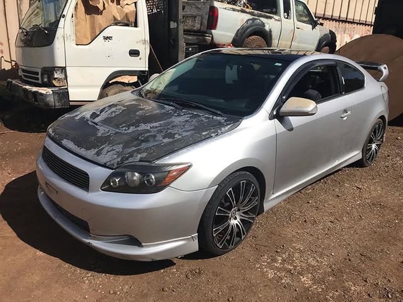 Silver Scion tC coupe with a damaged hood and blacked-out wheels, parked in a lot next to a white truck and other vehicles.