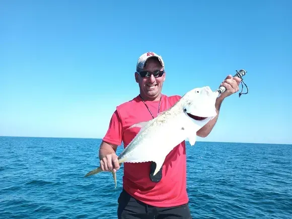 Man on boat holding up a large white fish, smiling. Blue sky and ocean in background.