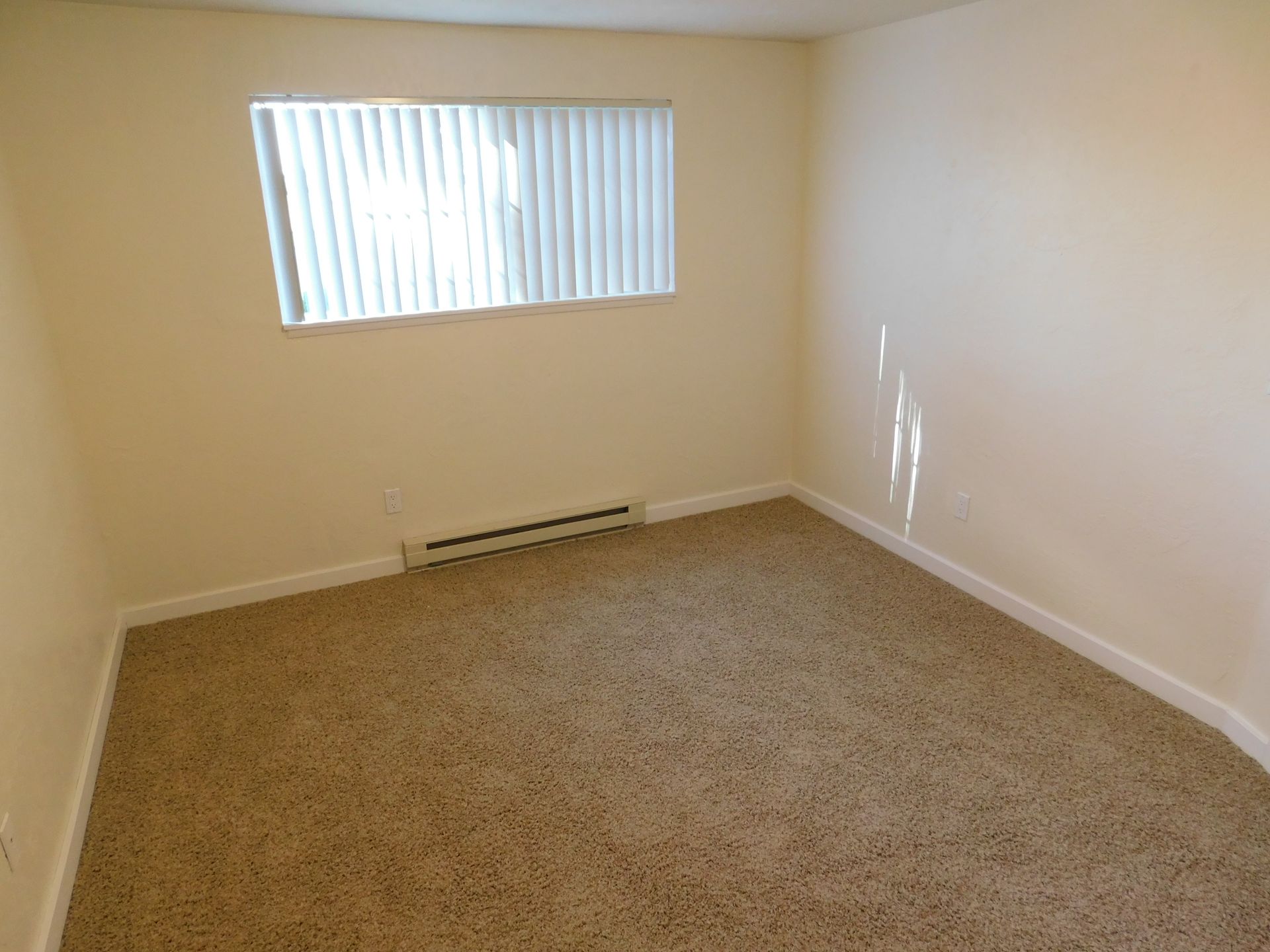 Empty room with tan carpet and walls; a window with blinds.