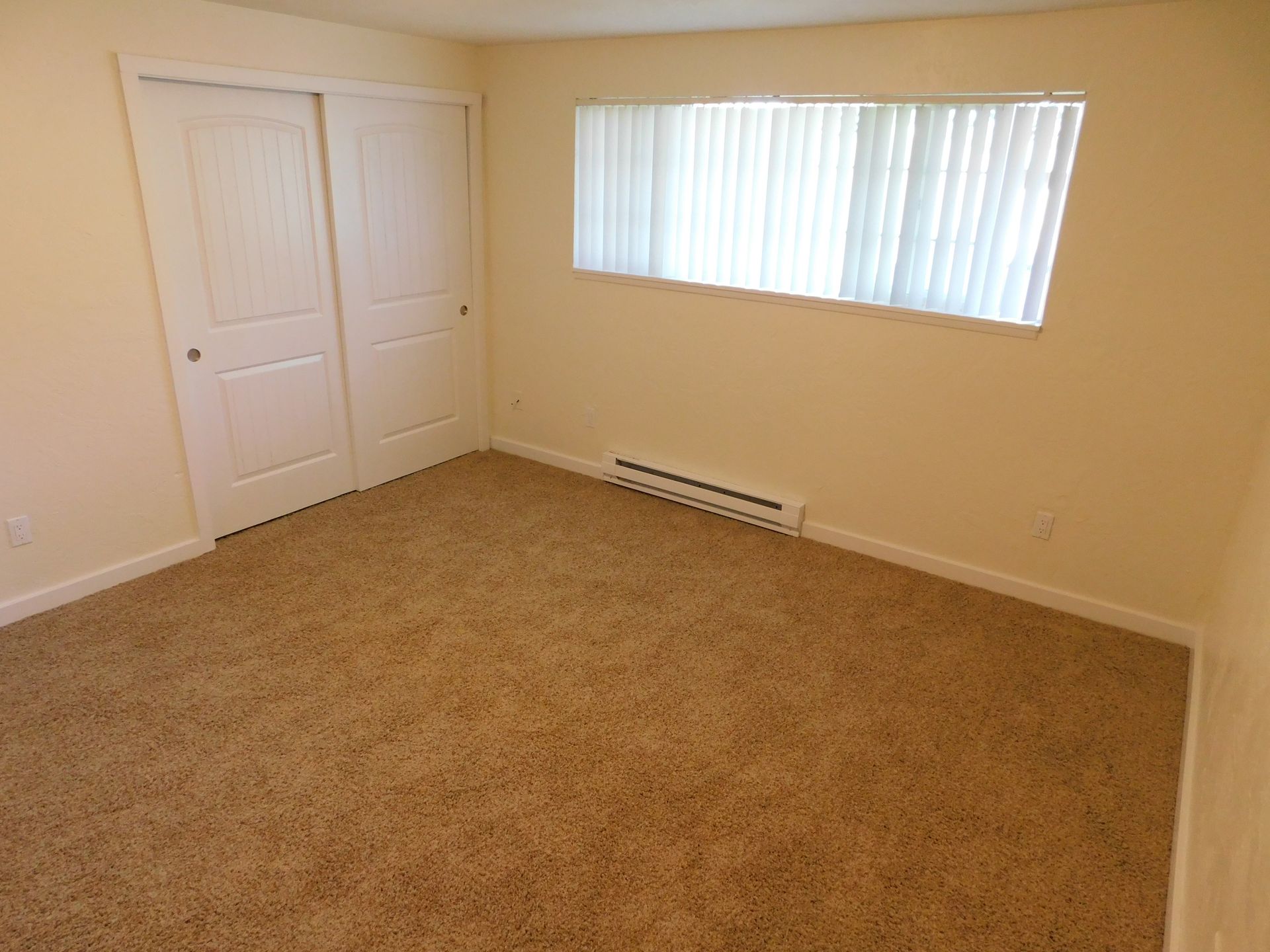 Empty beige bedroom with carpet, sliding closet doors, and a window with blinds.