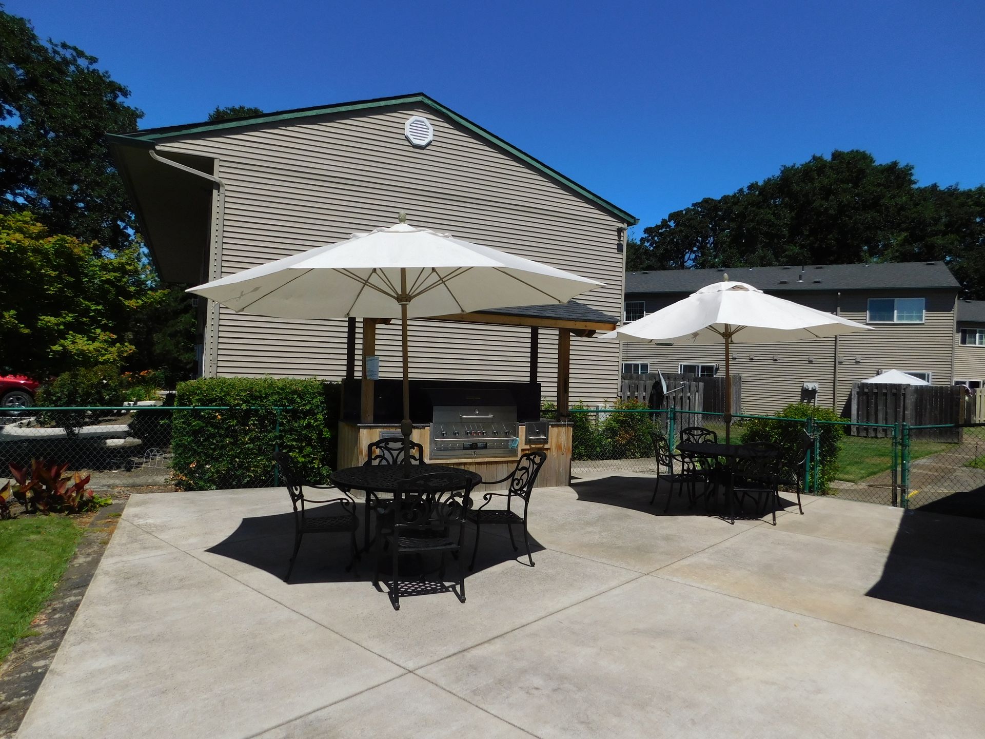Outdoor patio with umbrellas, tables, and a built-in grill, near an apartment building under a bright blue sky.