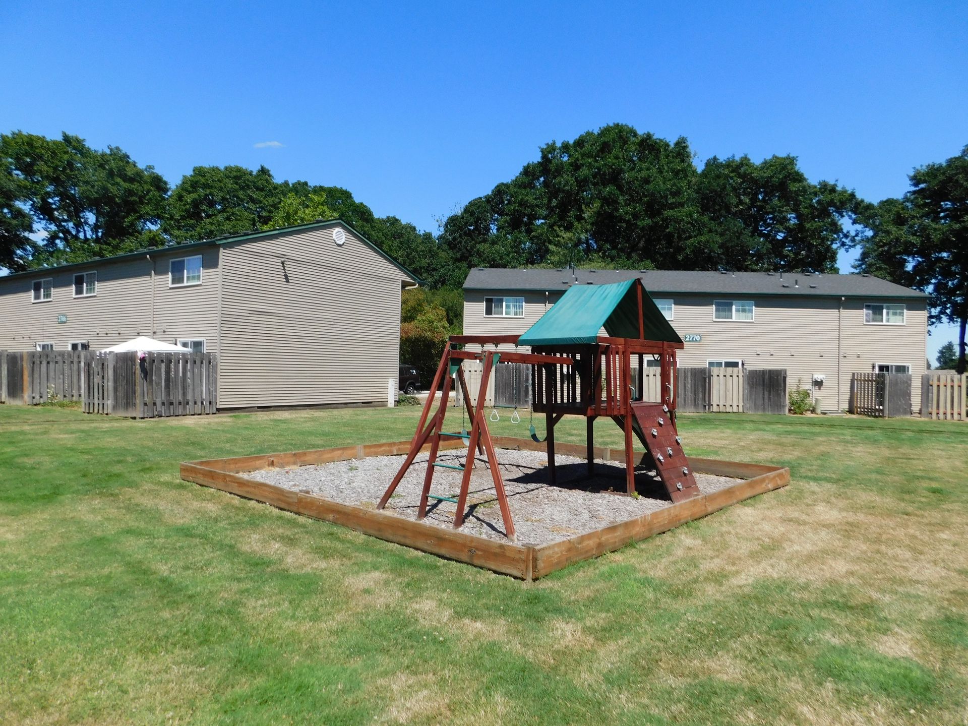 Playground with swings and slide surrounded by wood-framed gravel, in front of apartment buildings on a sunny day.