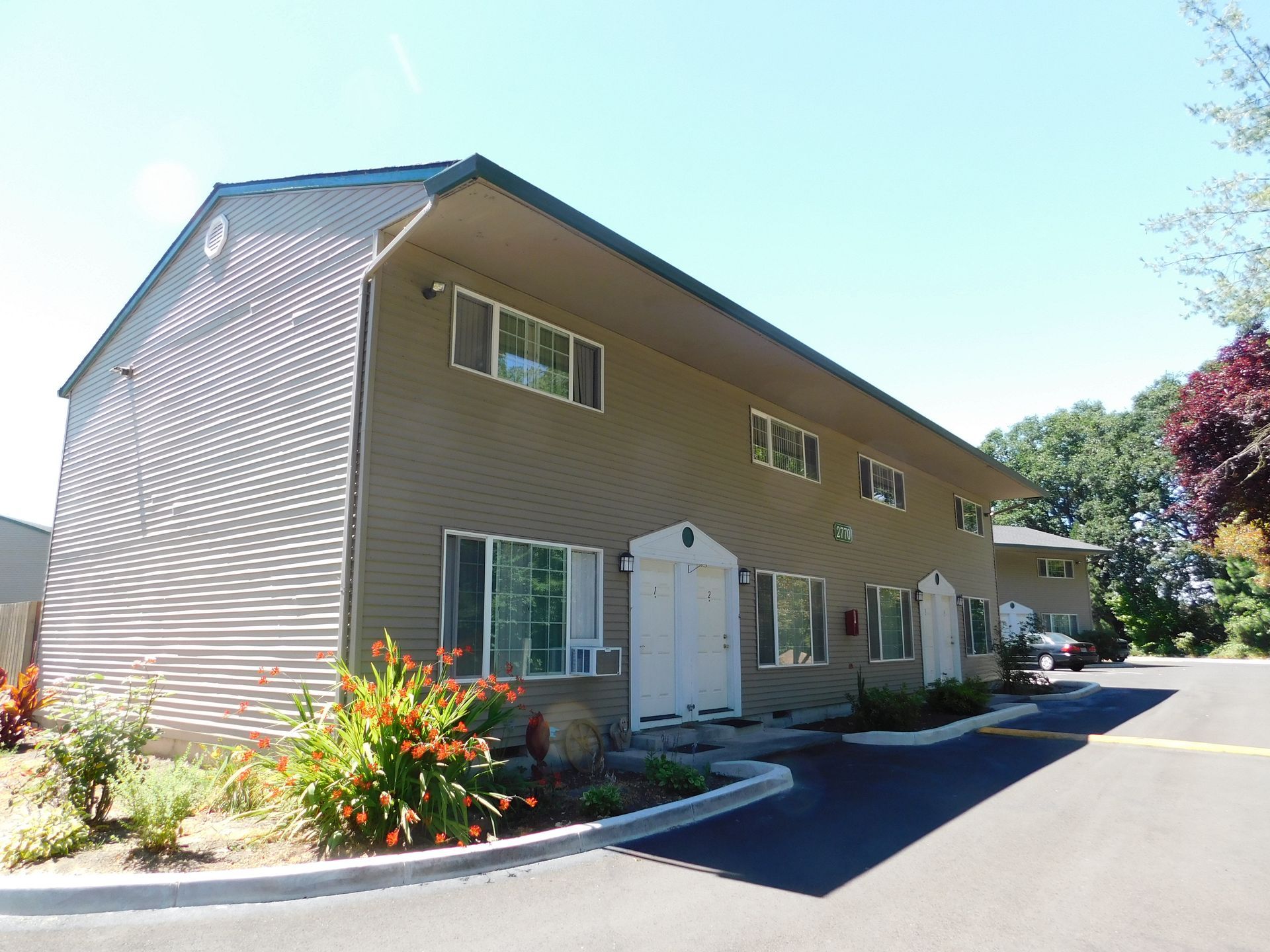 Two-story apartment building with tan siding and white doors, surrounded by landscaping.