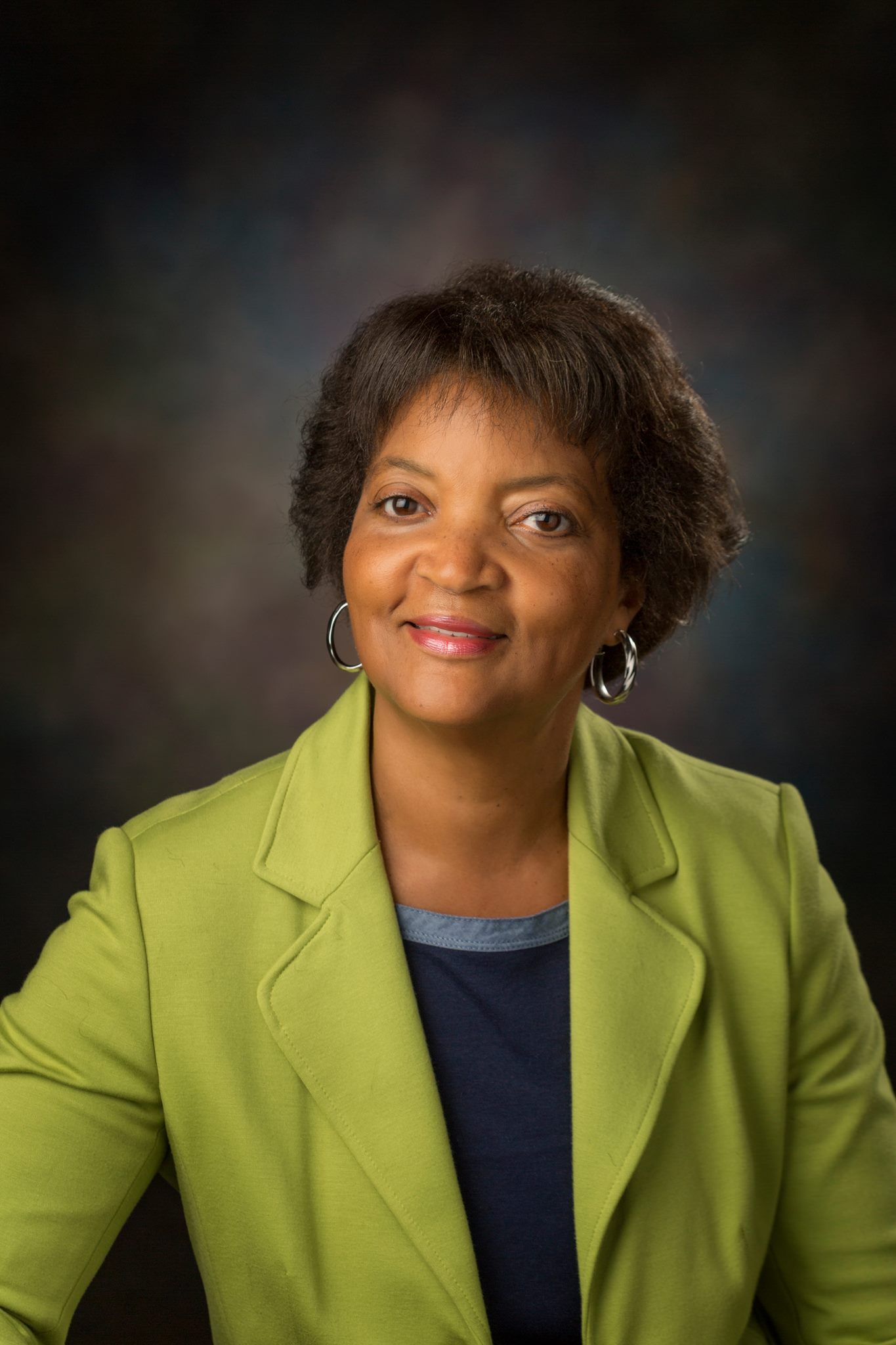 A woman wearing a green jacket and earrings is smiling for the camera.