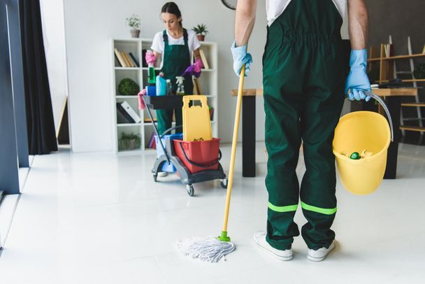 A man is holding a mop and a bucket while a woman is cleaning the floor.