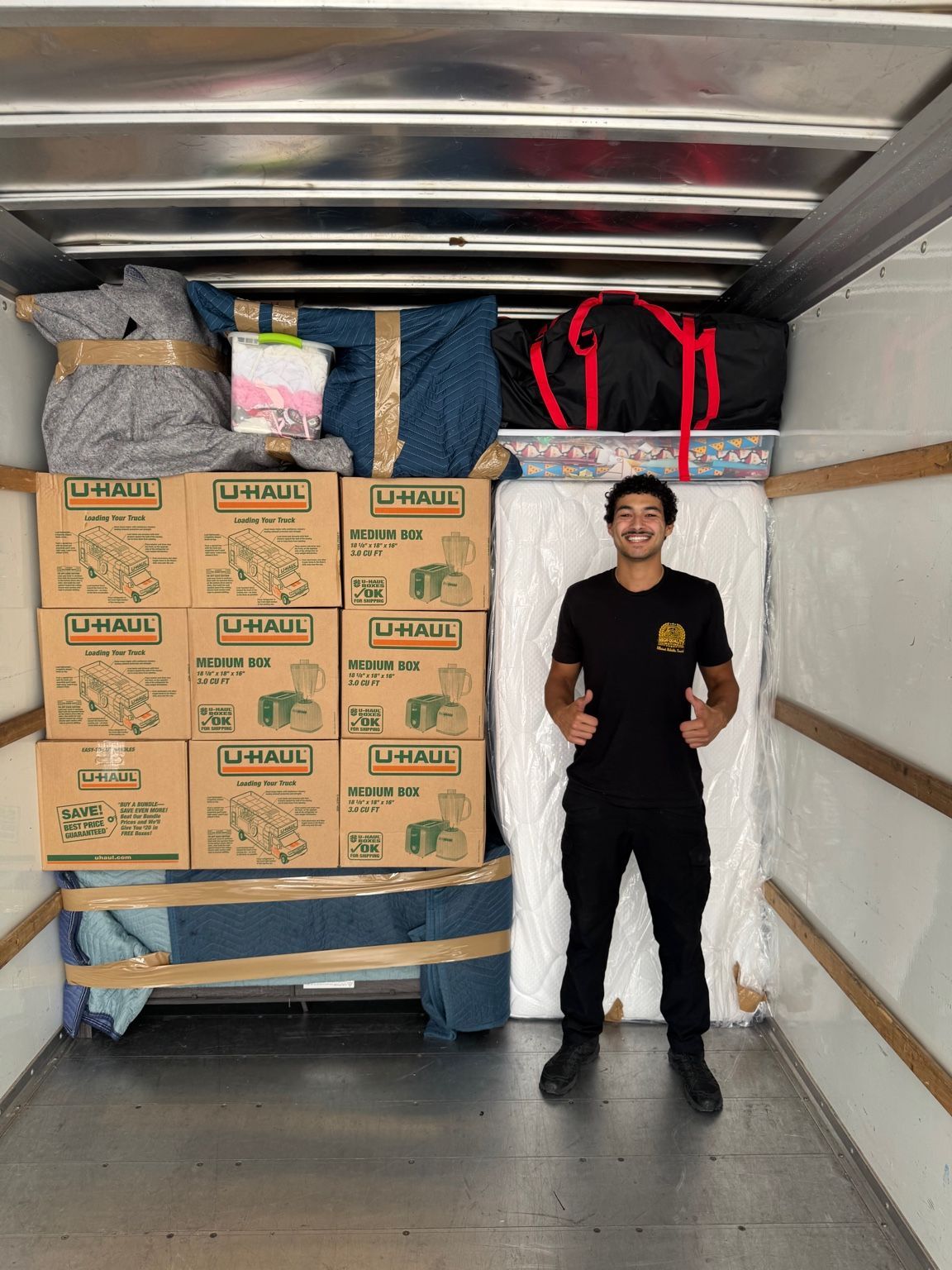 Man standing in a truck with thumbs up, surrounded by boxes, furniture, and luggage.