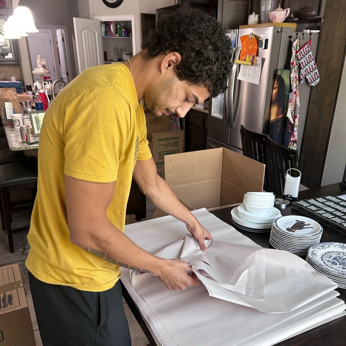 Man in yellow shirt wraps dishes in packing paper on a table. Cardboard box in background.