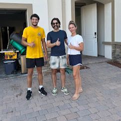 Three people give thumbs-up outside a house with moving supplies nearby.