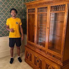 Man standing next to a large, ornate wooden cabinet. He wears a yellow shirt and black shorts.