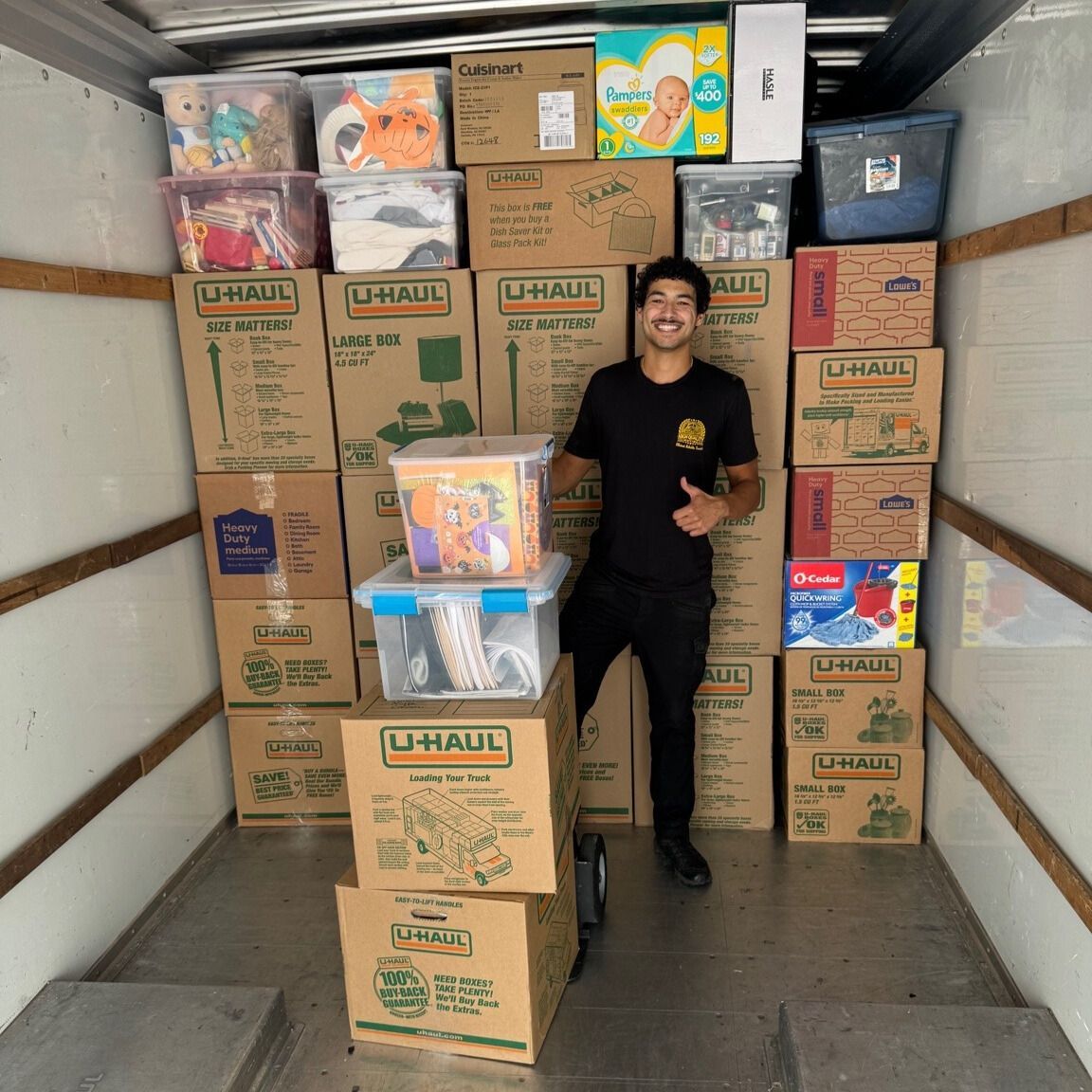 Man in black outfit gives thumbs up inside a moving truck stacked with U-Haul boxes and clear storage bins.