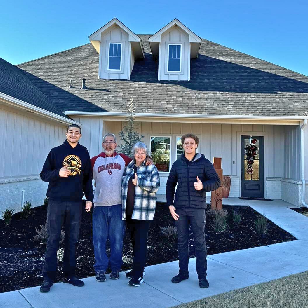 Three people give thumbs-up outside a house with moving supplies nearby.